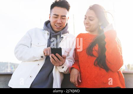 Multirassische Paare in legerer Kleidung surfen Internet auf Handy, während sie in der Nähe von Zaun und Fahrräder auf der sonnigen Straße der Stadt stehen Stockfoto