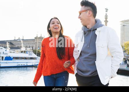 Glückliches multirassisches Paar, das sich mit einem Lächeln ansah, während es im Hafen nahe dem Meer mit einer festgetäuten Yacht in der Stadt spazieren ging Stockfoto