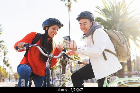 Positives multirassisches Paar in Helmen und Rucksäcken, das auf dem Handy surft, während es auf Fahrrädern auf einer sonnigen Sommerstraße mit grünen Bäumen sitzt Stockfoto