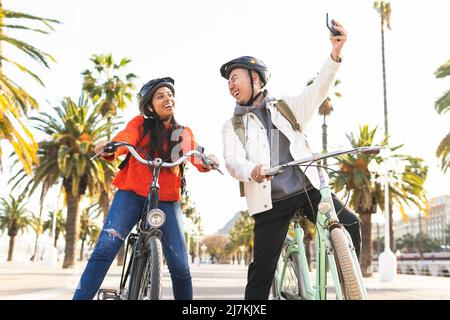 Von unten positives Paar in Helmen Fahrrad fahren auf Asphaltstraße und Selfie mit Smartphone in der Stadt mit grünen Bäumen am Sommertag Stockfoto