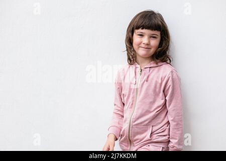 Positives kleines Mädchen in beiläufiger rosa Kleidung mit dunklem Haar, lächelnd und mit Blick auf die Kamera, während es auf der Straße gegen die weiße Wand steht Stockfoto