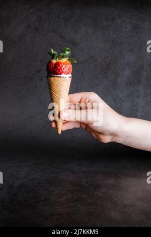 Erntehand unkennbare Person hält leckeren süßen Eiskegel mit reifen roten Erdbeeren vor grauem Hintergrund im Studio Stockfoto