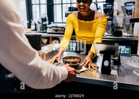 Glückliche afroamerikanische Kellnerin gab Tablett mit Smoothie-Schüssel und Löffel, die unerkennbaren Kunden über dem Tresen in einem kleinen Café servierte Stockfoto