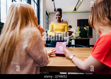 Eine ziemlich lächelnde, afroamerikanische junge Kellnerin nimmt Bestellungen von anonymen Freundinnen an, die am Tisch neben dem Fenster im Café sitzen und auf die Speisekarte zeigen Stockfoto