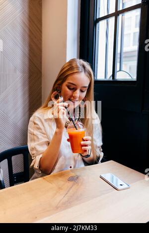 Schöne, fröhliche, moderne blonde Frau mit stilvollen Ohrringen, die Fruchtsaft genießen, während sie am Holztisch im Café sitzt Stockfoto