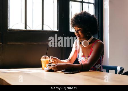 Schöne fröhliche junge afroamerikanische Frau, die Kaffee und Fruchtsaft in einem kleinen Café trinkt Stockfoto