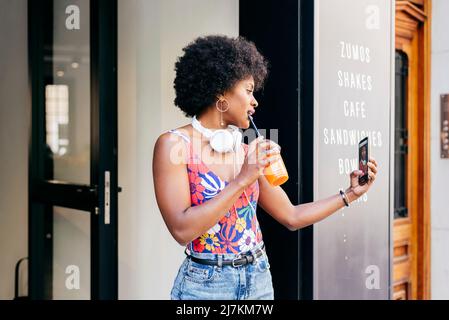 Seitenansicht einer ziemlich modernen afroamerikanischen Frau mit Kopfhörern am Hals und Orangensaft und einem Smartphone in den Händen, das den Coffee Shop verlässt Stockfoto