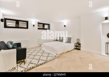 Komfortables weißes Bett mit Kissen auf Teppich und schwarzem Sessel im hellen, stilvollen Schlafzimmer mit kleinen Fenstern und glühender Lampe Stockfoto