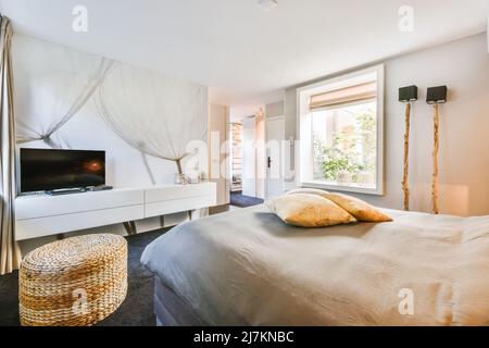 Modernes Fernsehgerät auf weißem Schrank vor einem bequemen weichen Bett im geräumigen Schlafzimmer mit Rattan-Hocker und kreativen Holzlampen Stockfoto