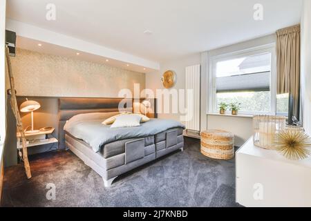 Komfortables Bett mit Kissen und Decke zwischen Nachttischen mit leuchtenden Lampen im stilvollen Schlafzimmer mit Fenster zu Hause platziert Stockfoto