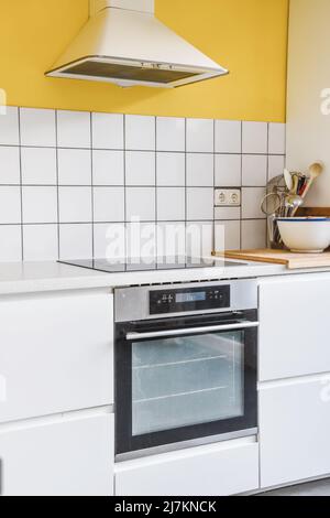 Weiße Schränke in hellen stilvollen Küche mit modernen eingebauten Backofen und Herd in geräumigen Wohnung mit gelber Wand platziert Stockfoto