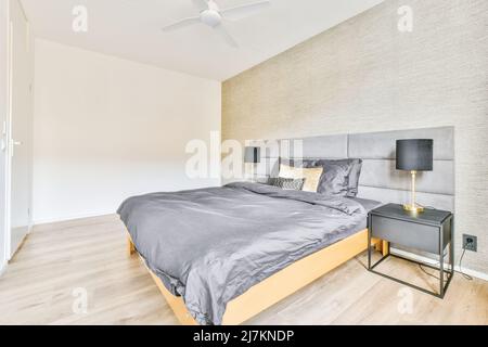 Komfortables Bett mit grauer Decke und Kissen zwischen Beistelltischen und Lampen im stilvollen Schlafzimmer zu Hause Stockfoto