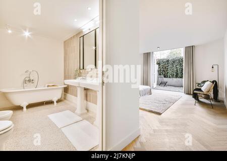 Interieur des eleganten Badezimmer mit weißer Badewanne und Waschbecken im geräumigen Schlafzimmer mit Parkettboden und Balkon in zeitgenössischem Haus Stockfoto