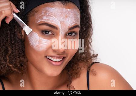 Selbstbewusstes lächelndes, junges, ethnisches Frauenmodell mit lockig dunklem Haar und Haarband, das mit Pinsel Maske auf das Gesicht aufgesetzt und die Kamera gegen das Whi angeschaut hat Stockfoto