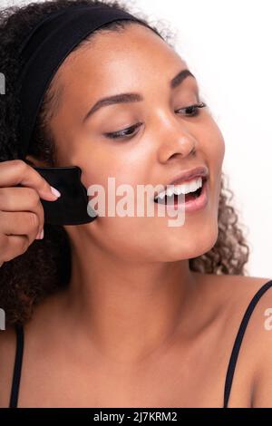 Optimistische junge hispanische Frau mit lockigen dunklen Haaren lächelt, während sie Gesichtsmassage mit einem Gua Sha-Werkzeug vor weißem Hintergrund macht Stockfoto