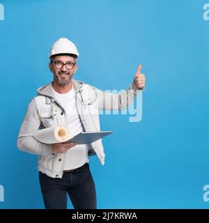 Ein hübscher Architekturmann mittleren Alters, der eine Rolle von Tapeten und einem digitalen Tablet-pc in einem trendigen legeren Denim-Outfit hält, einen Hut oder Helm mit dem Daumen nach oben oder perfekt isoliert auf blauem Hintergrund. Stockfoto
