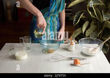 Crop anonyme Kochen in Schürze Gießen Öl in Glasschüssel mit Ei, während die Pfannkuchen am Tisch mit verschiedenen Zutaten Stockfoto