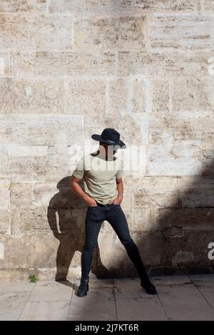 Ganzkörper von jungen trendigen Mann in stilvollen Hut in der Nähe verwitterte Steinmauer an sonnigen Sommertag in der Stadt stehen Stockfoto