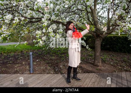 Moskau, Russland. 9.. Mai 2022. Eine Frau macht ein Selfie für Instagram vor dem Hintergrund blühender Obstbaumblumen und einer festlichen roten Flagge am Victory Day im Muzeon Park, Russland Stockfoto