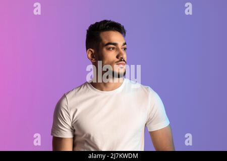 Ernsthafter junger arabischer Mann im weißen T-Shirt posiert und schaut in Neonlicht weg Stockfoto