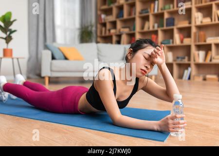 Erschöpfte asiatische Dame, die Schweiß von der Stirn abwischt, sich nach dem Training zu Hause müde fühlt und auf einer Yogamatte liegt Stockfoto