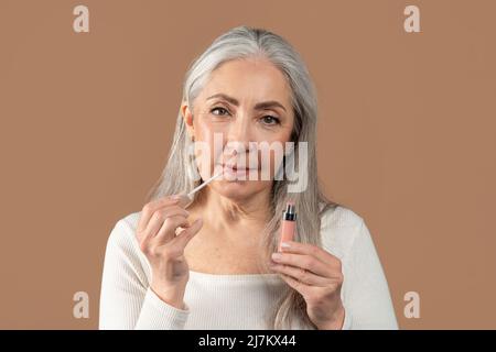 Schönheitsporträt der älteren Frau, die Lipgloss anwendet und die Kamera auf braunem Studiohintergrund anschaut. Organisches Make-up Stockfoto