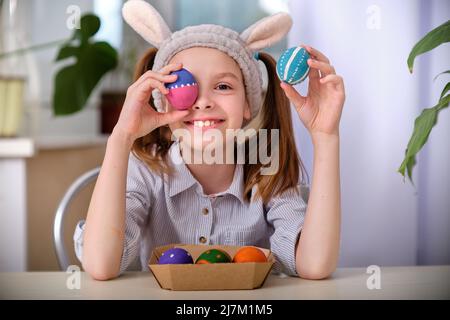 Ein positiv verspieltes Mädchen in Hasenohren hält in der heimischen Küche bunte Eier in den Händen. Frohe Ostern. Osterhintergrund. Stockfoto