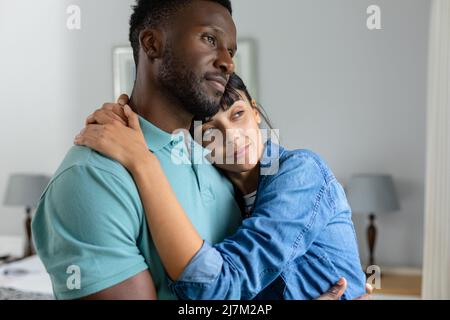 Kontemplatives, multirassisches junges Paar, das sich umarmt, während es zu Hause zusammensteht Stockfoto