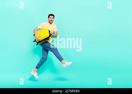 Ganzkörper-Foto von beeindruckt Brunet Kerl laufen mit Tasche tragen T-Shirt Jeans Sneakers isoliert auf blauem Hintergrund Stockfoto