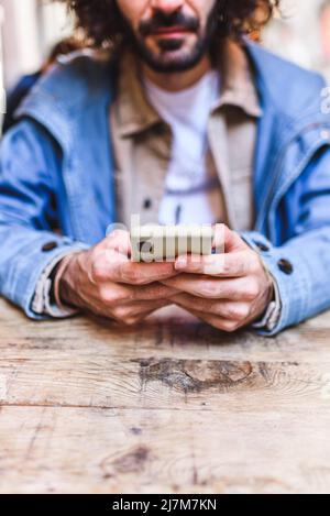 Weicher Fokus des bärtigen Mannes in legerer Kleidung, der SMS-Nachrichten auf dem Mobiltelefon sendet, während er am Holztisch im Restaurant sitzt Stockfoto