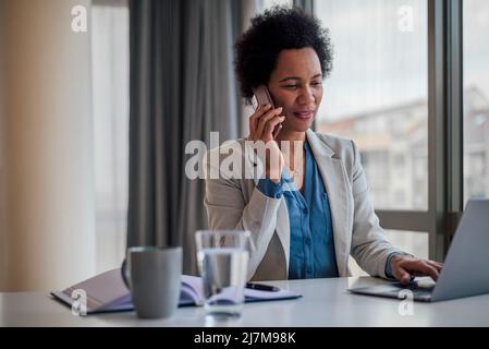 Geschäftsfrau im Gespräch auf dem Smartphone. Die Managerin arbeitet am Laptop, während sie am Schreibtisch sitzt. Sie ist in ihren Formalien in der Unternehmenszentrale. Stockfoto