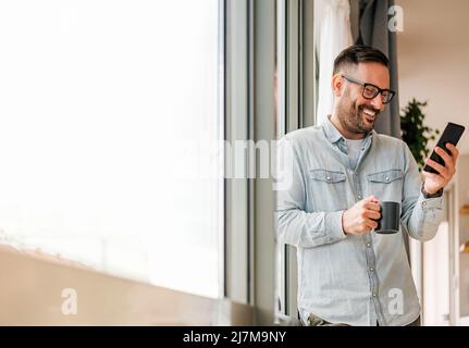 Lächelnder Mann Geschäftsmann in Casuals im Büro neben dem Fenster stehend mit Handy trinken Kaffee halten Kaffeetasse Kleinunternehmer Stockfoto