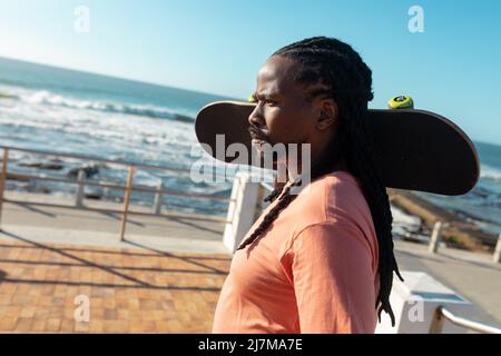 afroamerikanischer Mann, der an sonnigen Tagen an der Promenade Skateboard auf der Schulter trägt Stockfoto