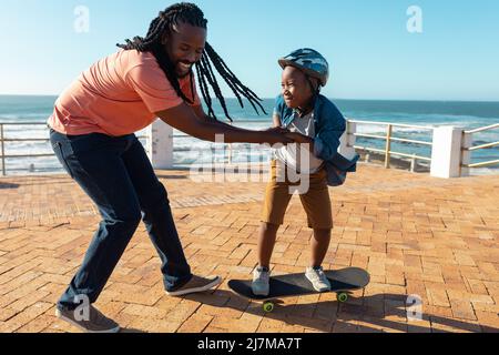 Fröhlicher afroamerikanischer Vater hilft Sohn beim Skateboarden auf der Promenade an sonnigen Tagen Stockfoto