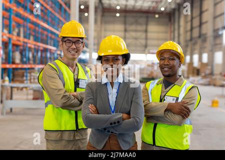 Lächelnde, multirassische weibliche und männliche Mitarbeiter tragen Hardhüte mit gekreuzten Armen im Lager Stockfoto
