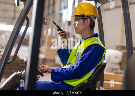 Seitenansicht eines reifen asiatischen Lagerarbeiters, der während der Fahrt mit dem Gabelstapler über Walkie-Talkie spricht Stockfoto