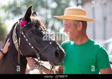 Ein älterer Mann in einem Strohhut lächelt ein Pferd an. Stockfoto