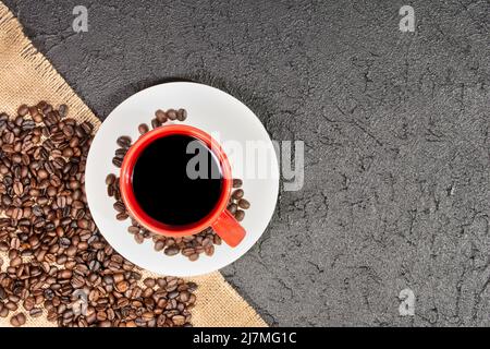 Blick von oben auf schwarzen Kaffee, der in einer roten Tasse auf einem weißen Teller serviert wird, umgeben von gerösteten Kaffeebohnen, die aus einem Sacksack auf einem dunklen strukturierten Backgr verschüttet werden Stockfoto
