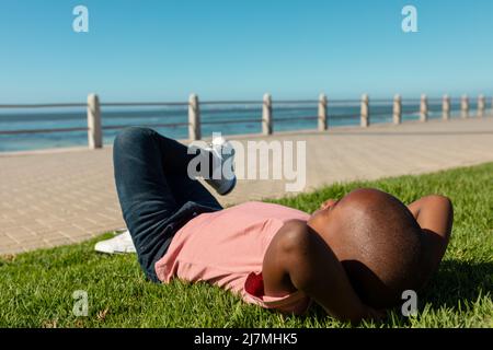 Die ganze Länge des afroamerikanischen Jungen entspannt, während er auf Gras an der Promenade während des sonnigen Tages liegt Stockfoto