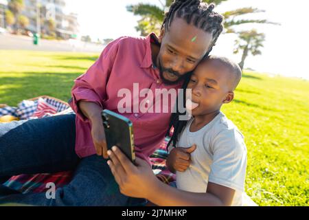 afroamerikanischer Junge ragt aus der Zunge, während er am sonnigen Tag ein Selfie mit dem Vater im Park nimmt Stockfoto