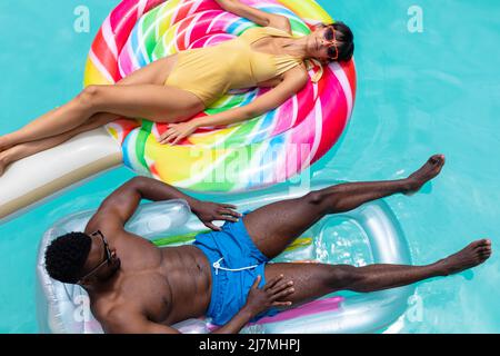 Overhead-Ansicht von multirassischen jungen Paar entspannen auf aufblasbaren Ringen im Schwimmbad. Unverändert, Zweisamkeit, Lebensstil, Freizeit, Bindung, Wochenende Stockfoto