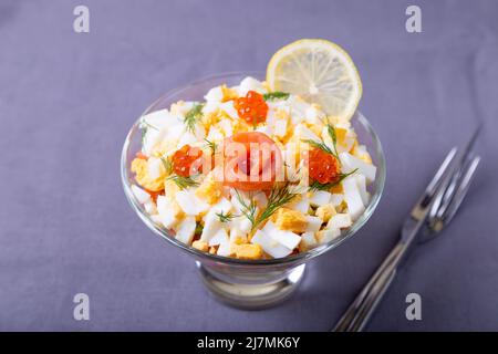 Geschichteter Salat mit gesalzener Forelle (Lachs), Kaviar, Eiern und Gemüse. Traditionelles Gericht, eine Portion in einem Glaskelch auf grauem Hintergrund. Nahaufnahme, Stockfoto