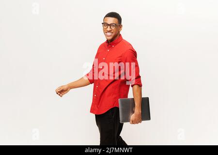 Fröhlicher, gutaussehender, afroamerikanischer junger Mann in rotem Hemd, der Laptop trägt und isoliert auf Weiß läuft. Freundliche, gut aussehende multirassische Studentin, Freiberufler in Brille Stockfoto