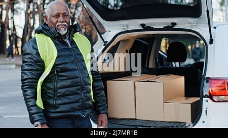 Älterer afroamerikanischer männlicher Kurier in Uniform nimmt Waren im Freien mit, reifer liefernder Arbeiter entlädt Kartons vom Autohandyman-Lader Stockfoto
