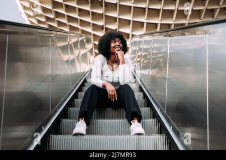 Lächelnde und nachdenkliche junge, schwarze Frau im Teenageralter mit afro-Haaren, die auf einer Rolltreppe im Freien sitzt. Stockfoto