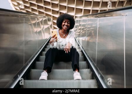 Lächelnde junge schwarze Teenagerin mit Afro-Haaren sitzt auf einer Rolltreppe im Freien und nimmt ein Selfie auf. Stockfoto