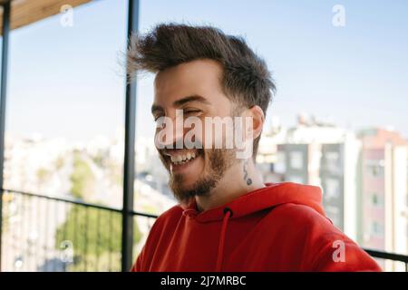 Fröhlicher junger Mann mit rotem Hoodie, lachend Nahaufnahme Porträt vor Stadthintergrund. Stockfoto