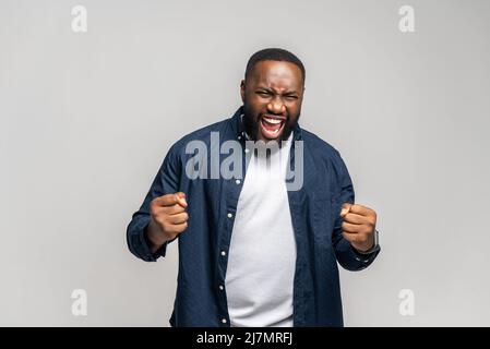 Gewinner-Konzept. Aufgeregter junger afroamerikanischer Mann, der ja schreit, glücklicher schwarzer Kerl, der geballte Fäuste in einer Siegesgeste hochhebt, isoliert auf einer grauen Wand Stockfoto