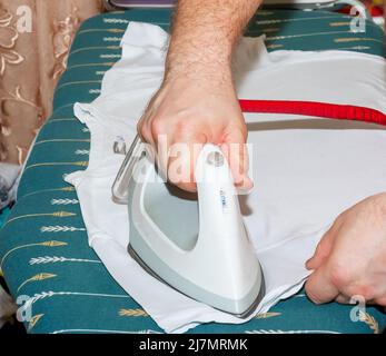 Ein Mann bügelt mit einem weißen Bügeleisen die Kleidung auf einem Bügelbrett. Hausarbeit und Haushaltskonzept. Bügeln von Kleidung zu Hause. Stockfoto