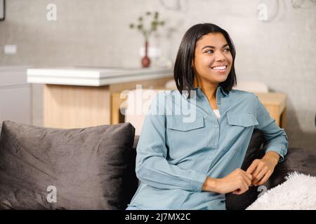 Nahaufnahme Porträt junge multirassische Frau in zu Hause Innenraum. Charmante Teenager-Dame, die wegschaut und lächelt, während sie sich allein ausruht. Heitere Frau, die ein Wochenende zu Hause verbringt Stockfoto